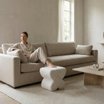Load image into Gallery viewer, Woman sitting on a beige sofa in a minimalistic living room with a wooden coffee table and white rug.